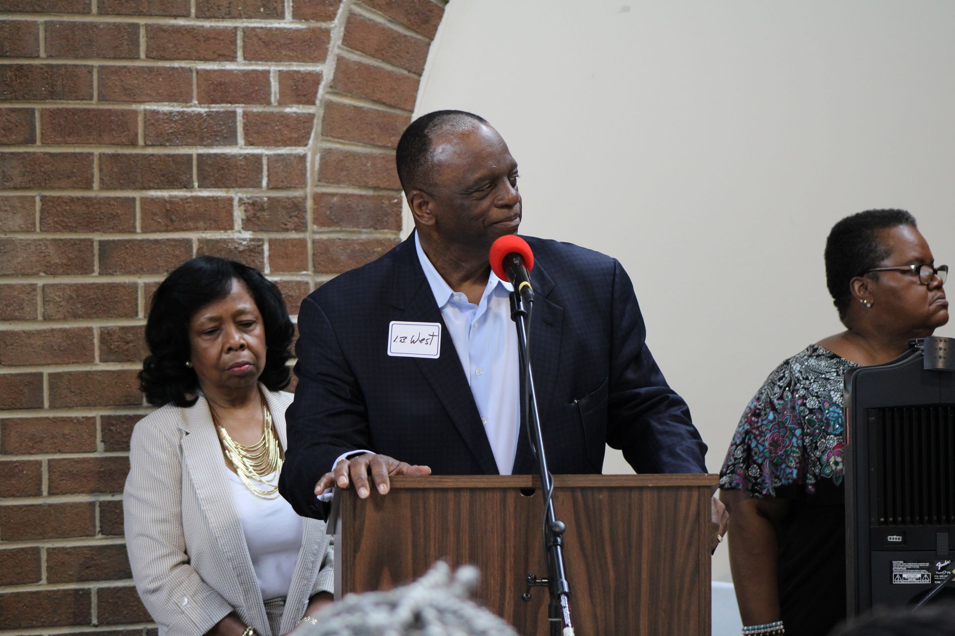 Man speaking at a podium with two women beside him, brick wall background.