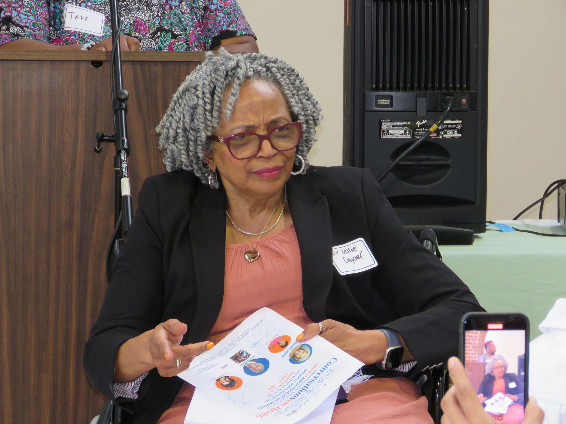 Woman in glasses holding paper, gesturing. Next to speaker, taking photo with phone.