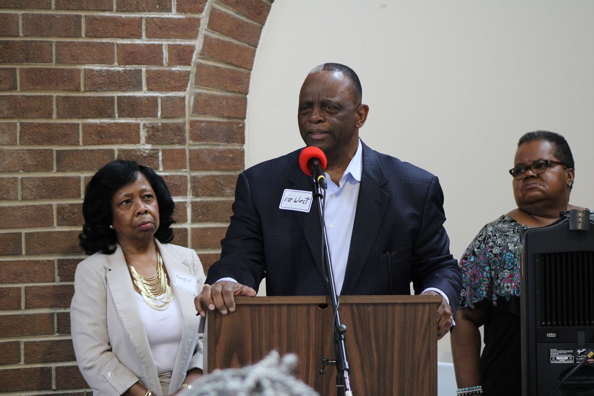 Man speaking at podium, flanked by two women, in front of a brick wall and speakers.