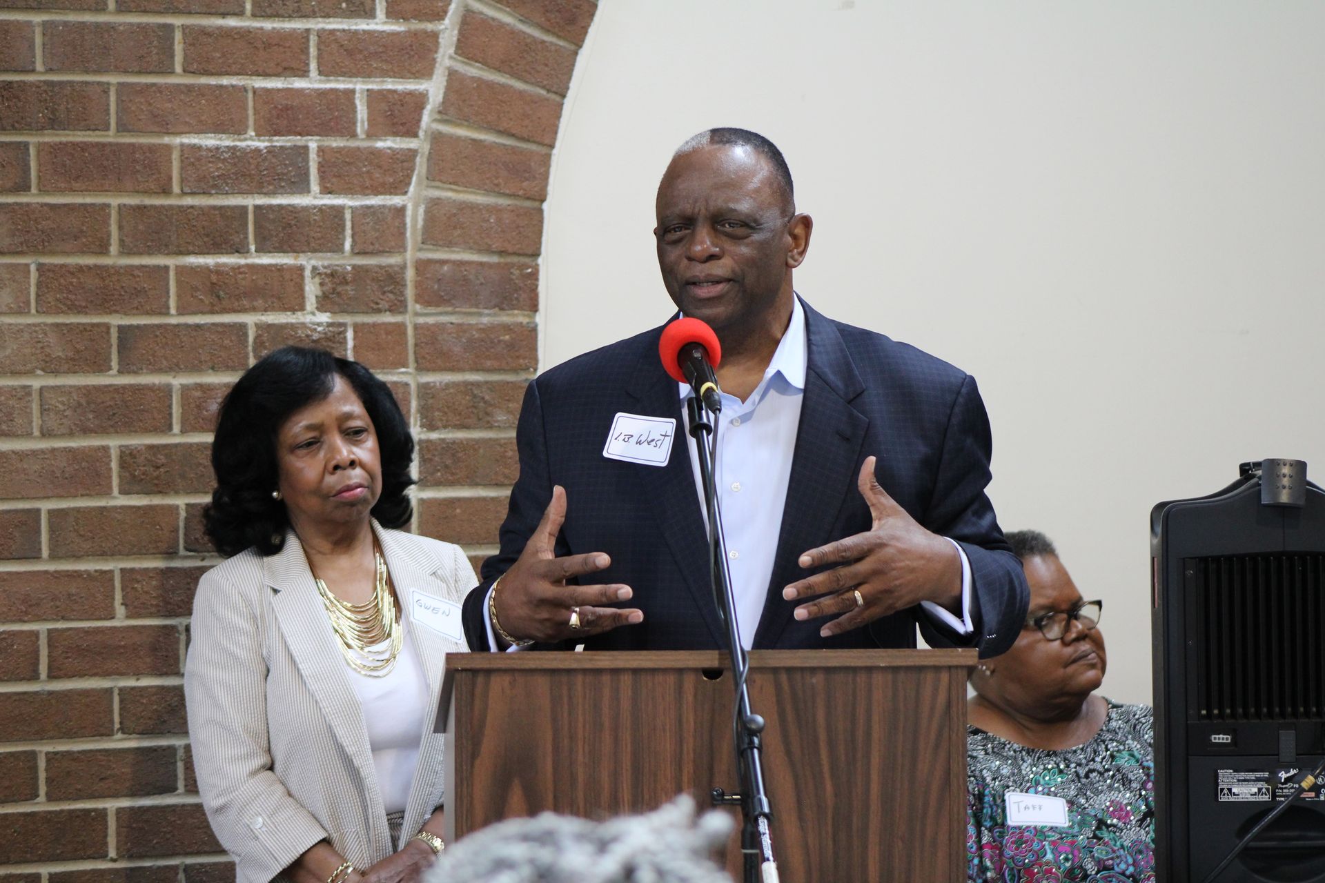Man speaking at a podium, gesturing. Woman stands to the side. Brick wall background.