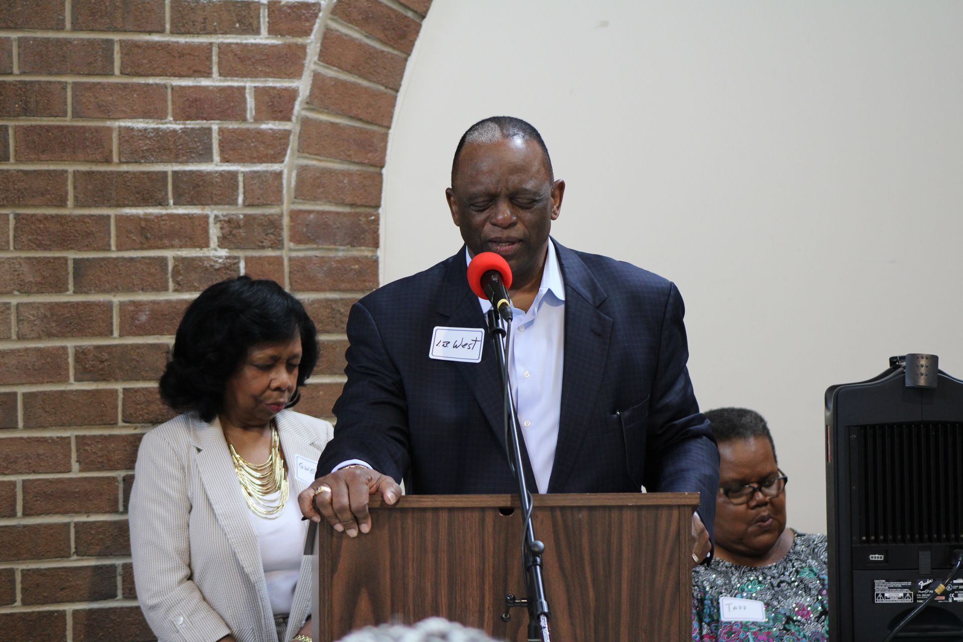 Man speaking at a podium with two women beside him, likely at an event. Brick wall background.