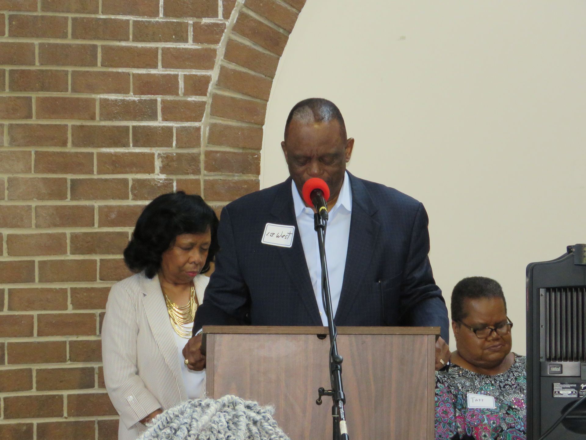 Man speaking at a podium with two women beside him, brick wall background.
