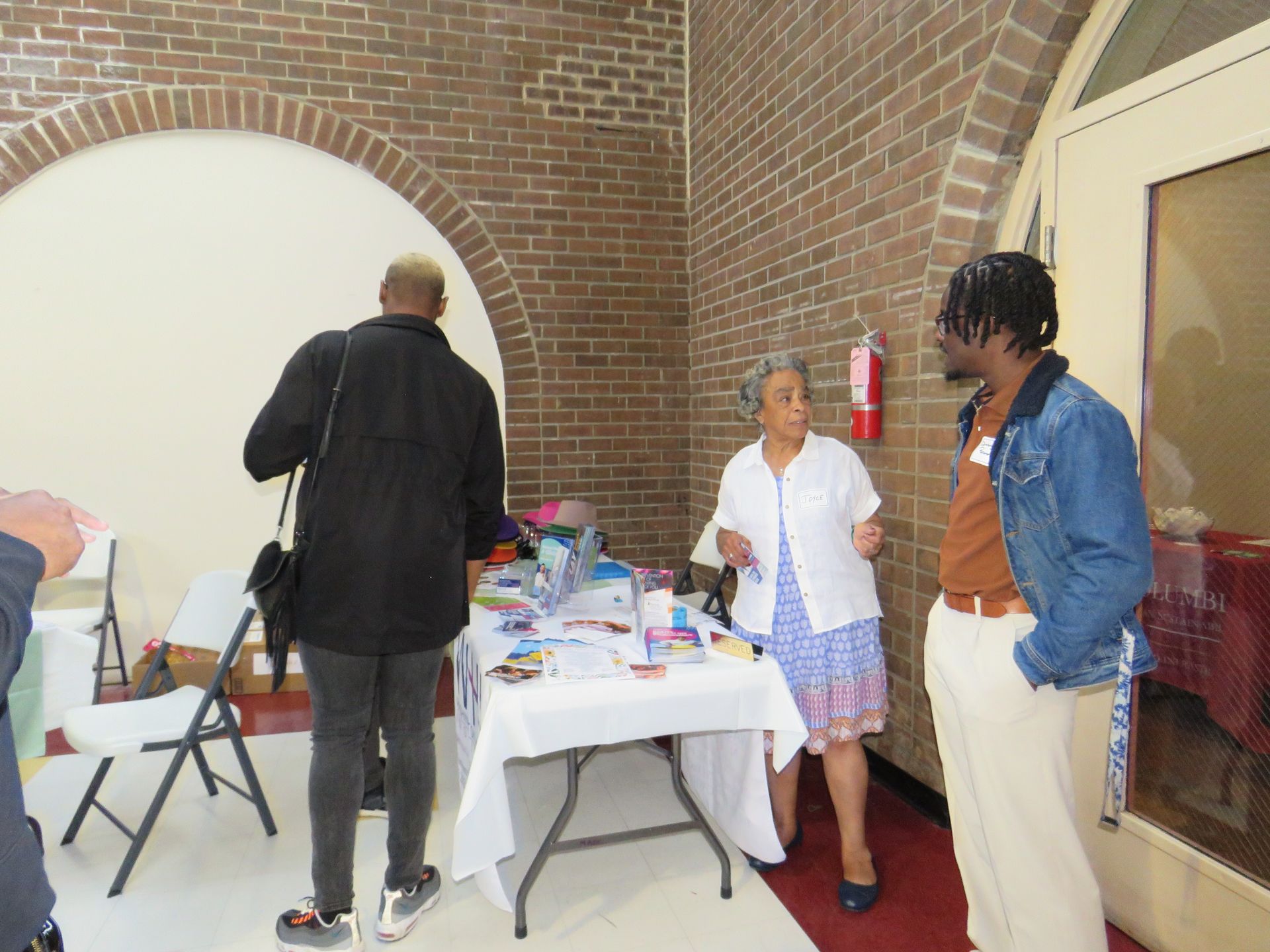 Three people at a table, inside a brick building. Two are talking, one is looking at items.