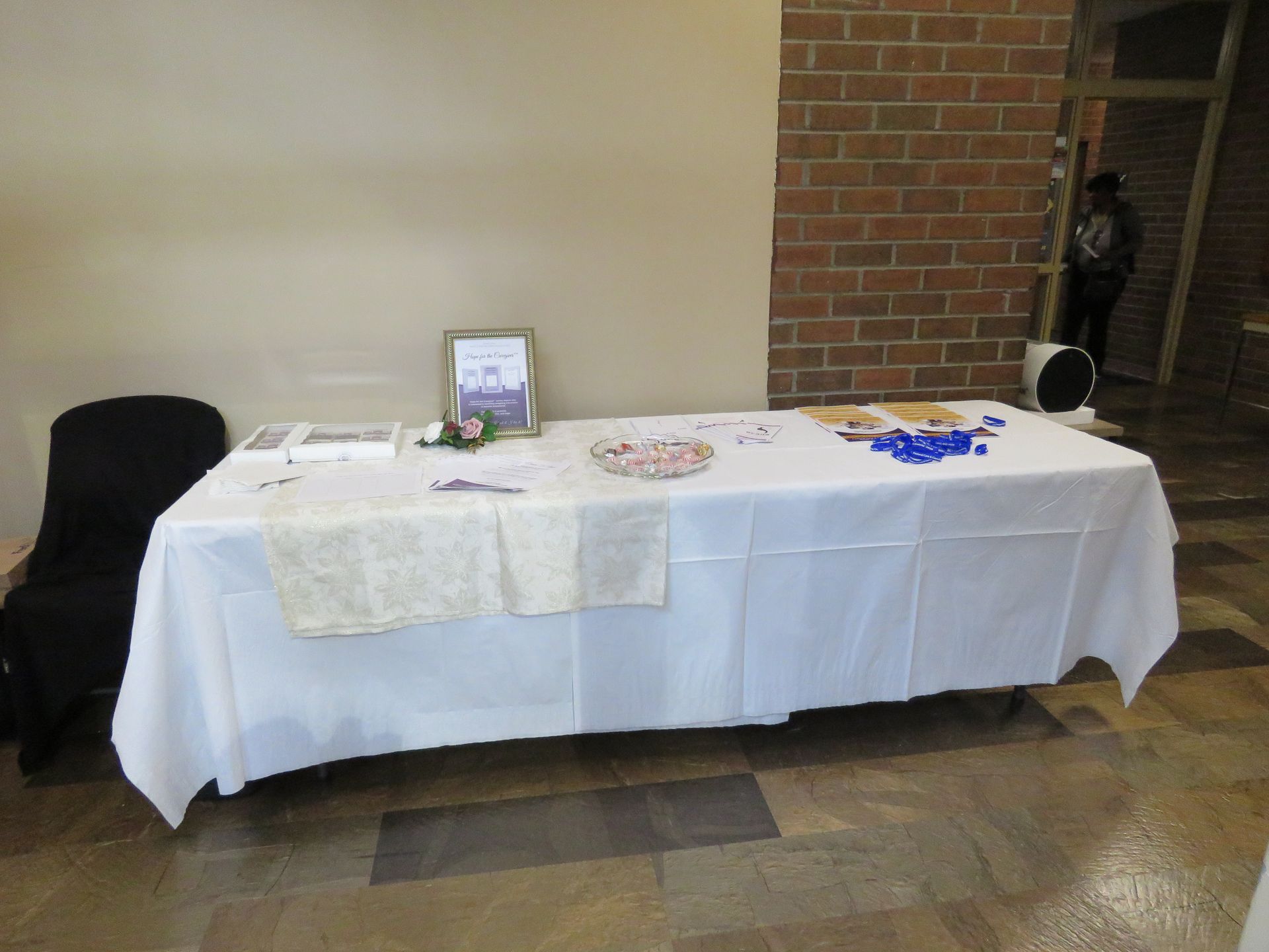 Table with white cloth, brochures, and candies. Black chair on left. Brick wall in background.