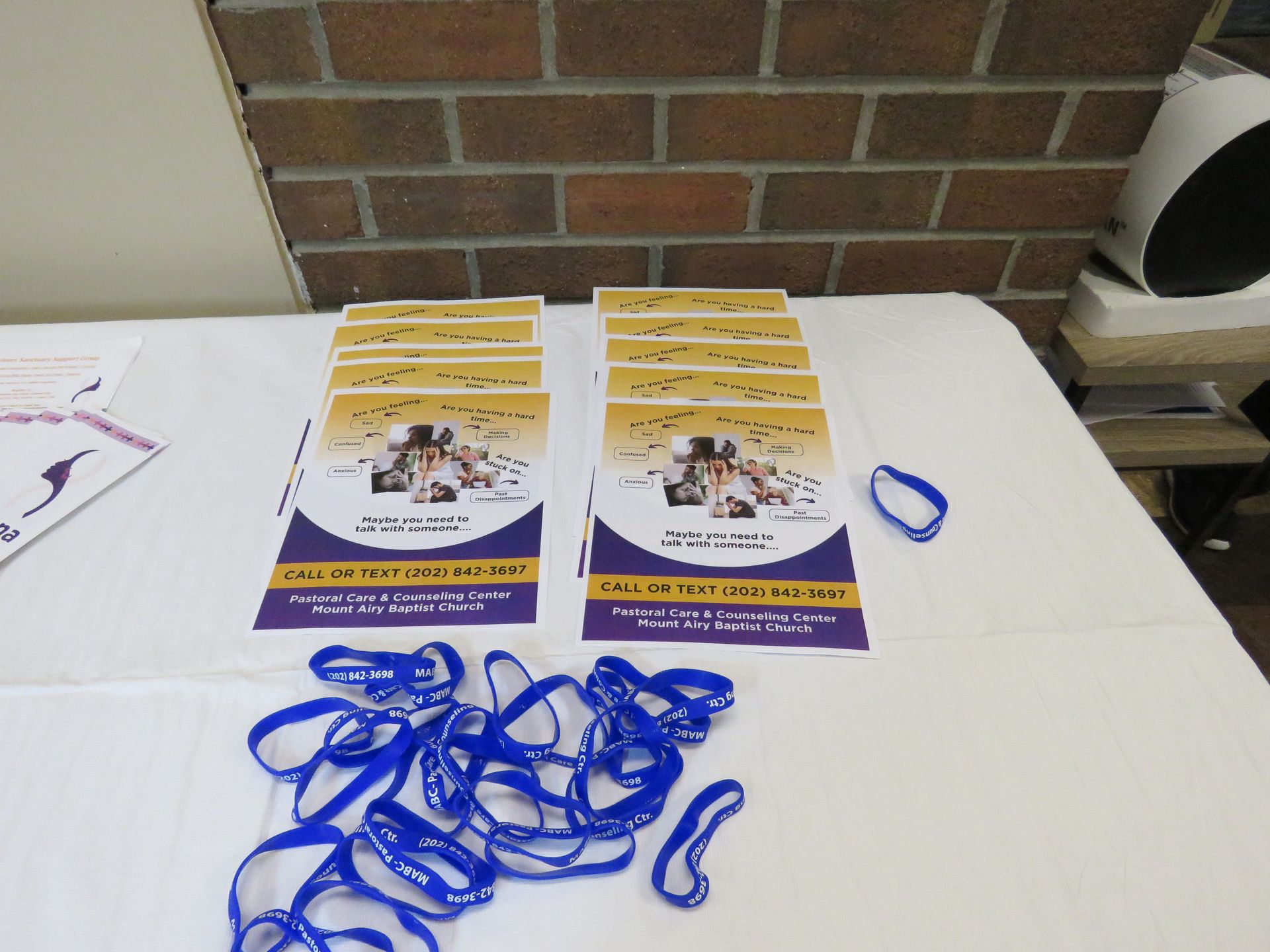 Table with stacks of purple/gold flyers and blue wristbands. Brick wall background.