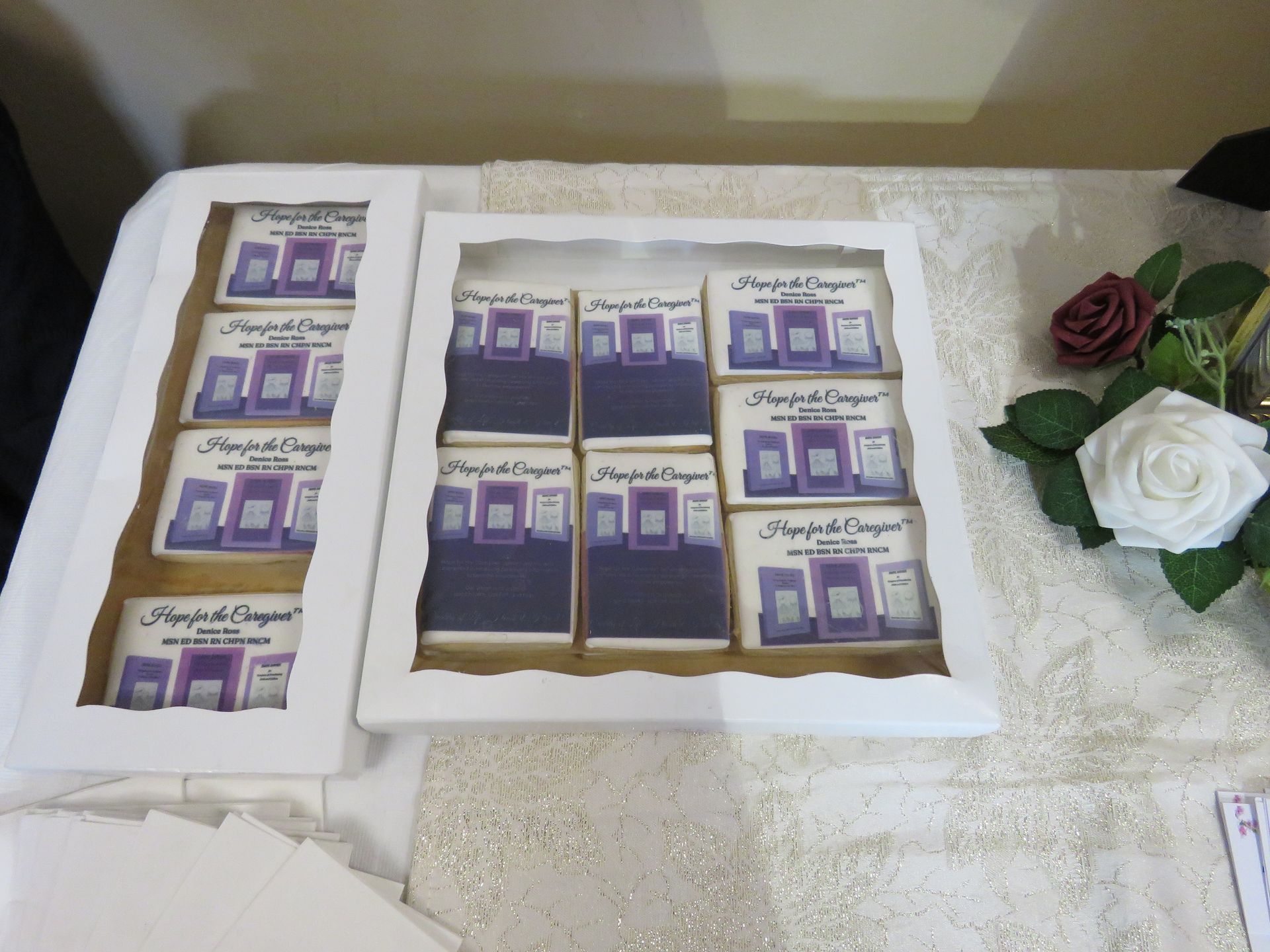 Cookies arranged in boxes, each decorated with a purple and white design. Roses sit nearby on a white tablecloth.