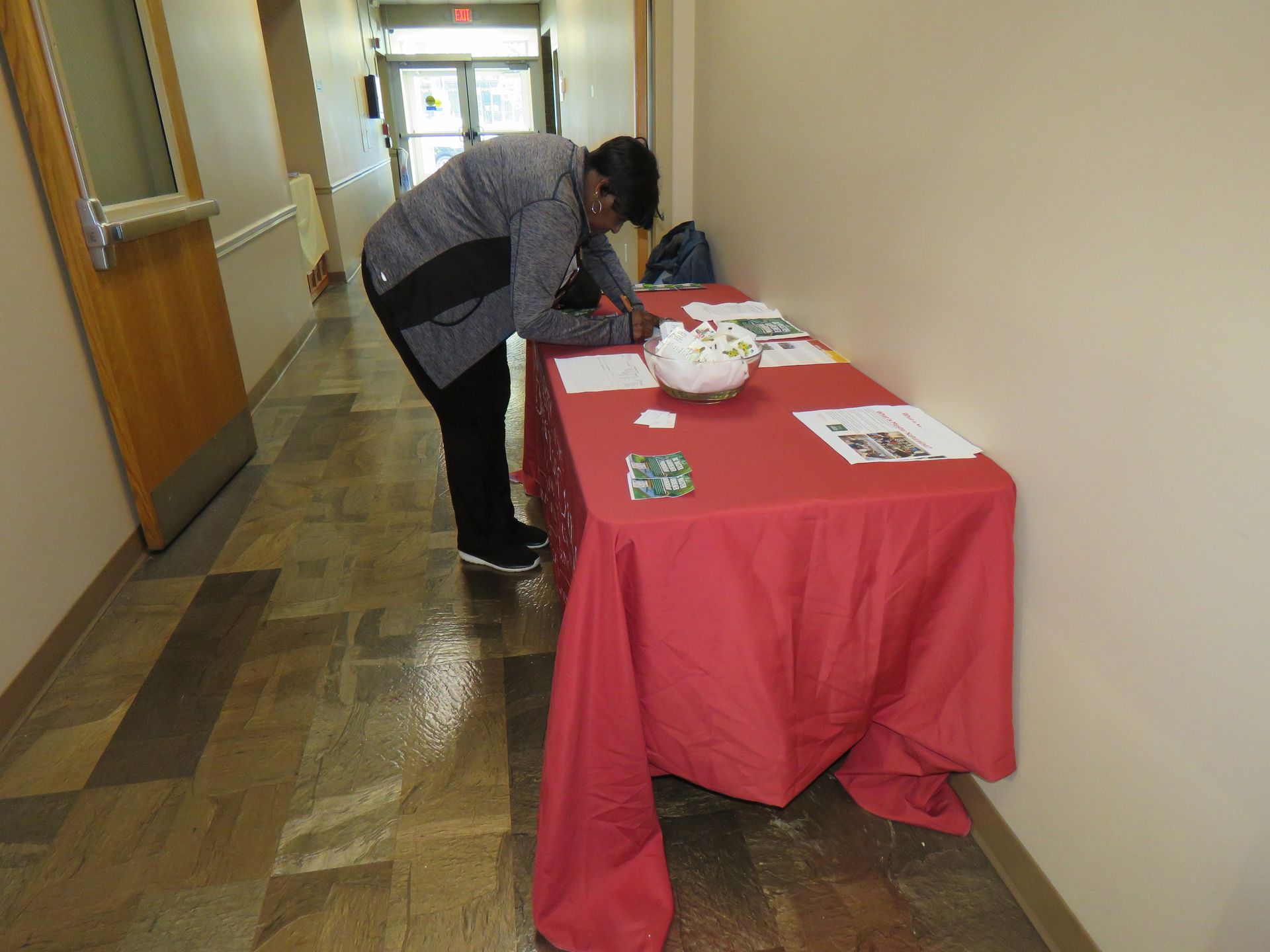 Person at a red table in a hallway, writing on papers. The table has items and a bowl.
