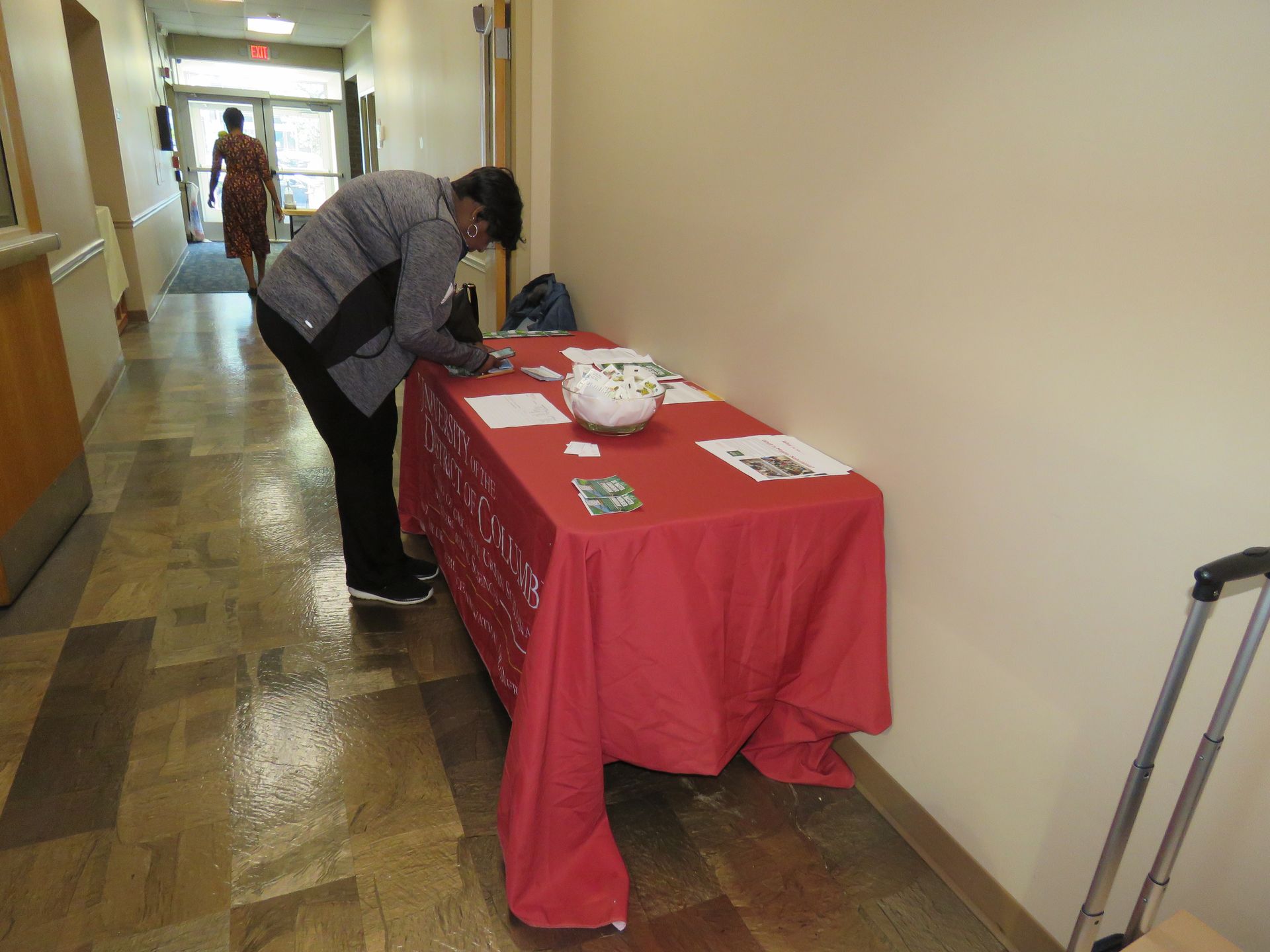 Person signing papers at a red-covered table in a hallway. A luggage cart is to the right.