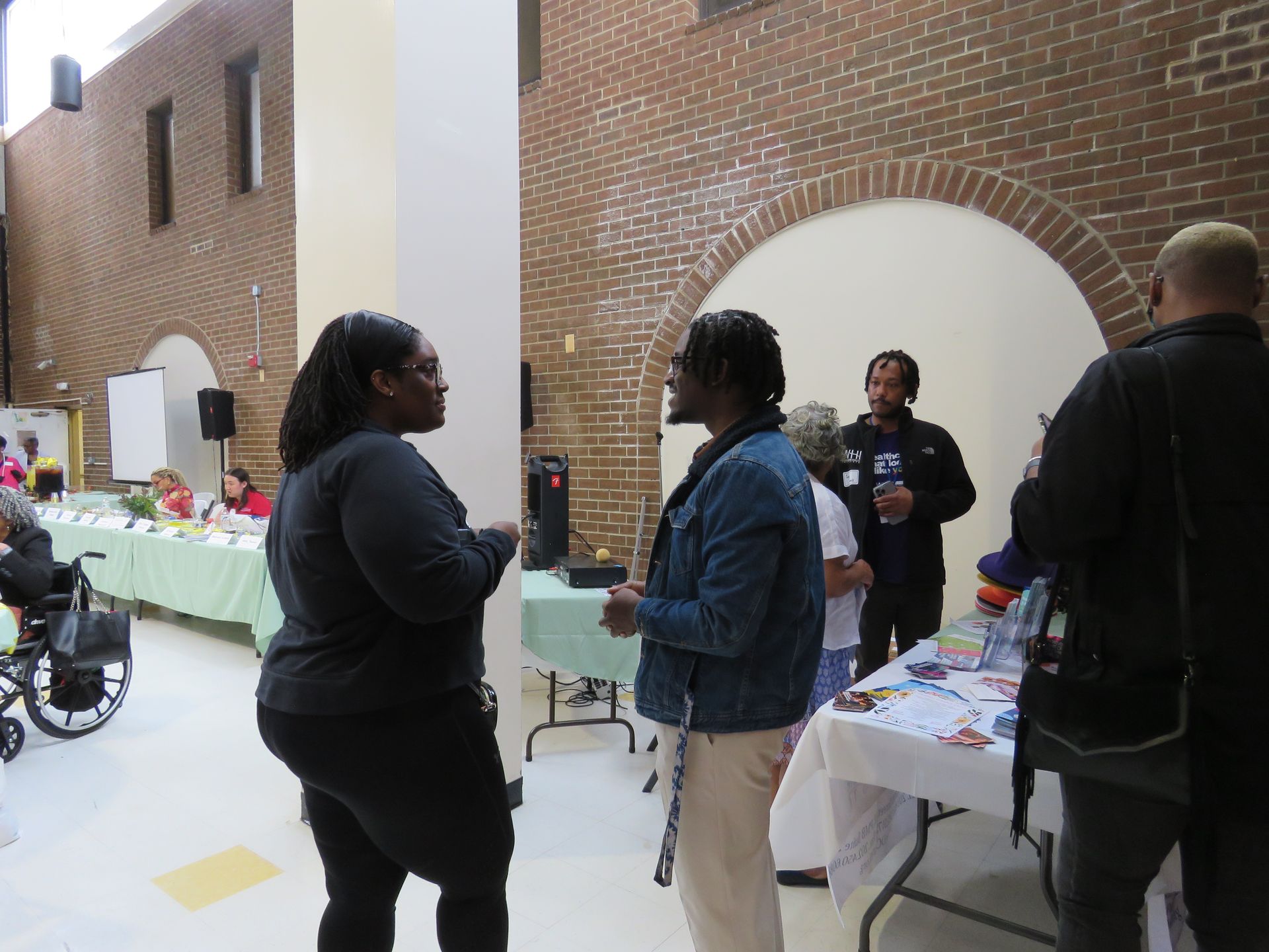 People at an event, talking and interacting near tables with information. Brick and archway background.