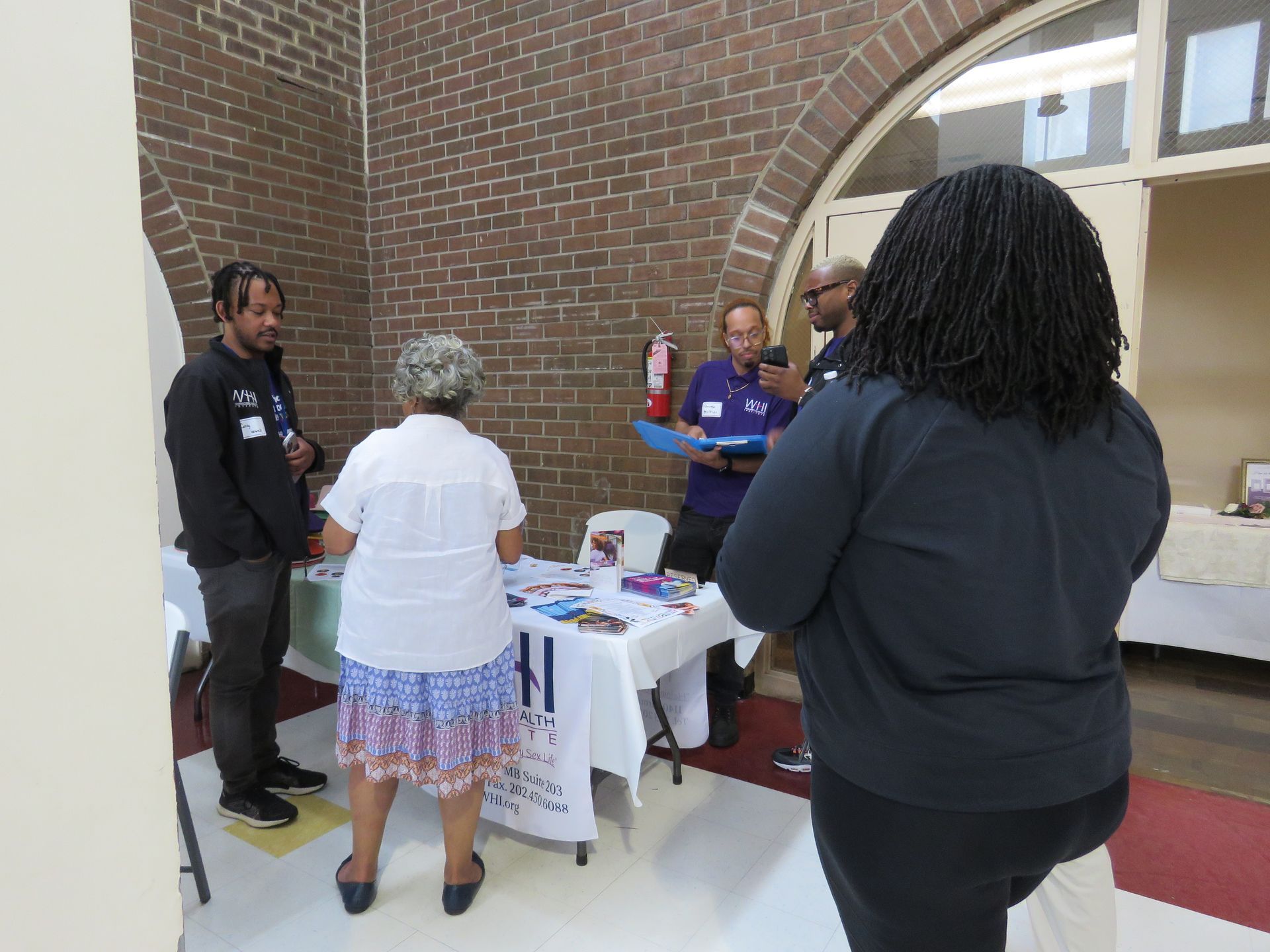 People at a table with brochures in a room with a brick wall. A fire extinguisher is visible.