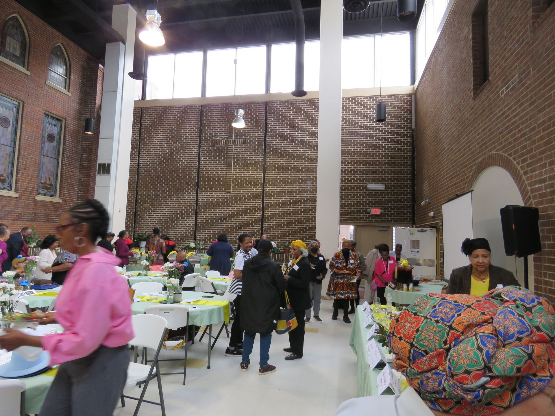 People gathered inside a building with tables, brick walls, and tall windows.