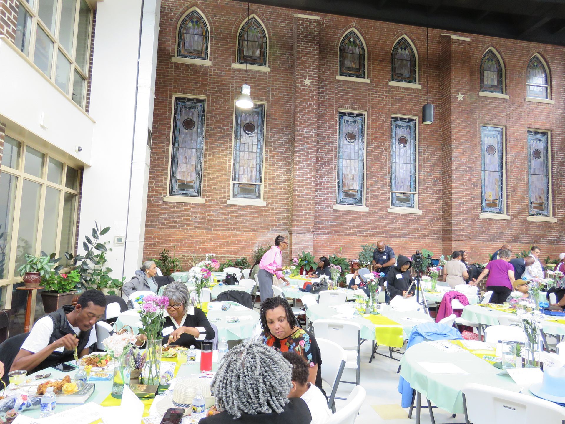 People eating at tables in a brightly lit room with large stained-glass windows on the brick wall.