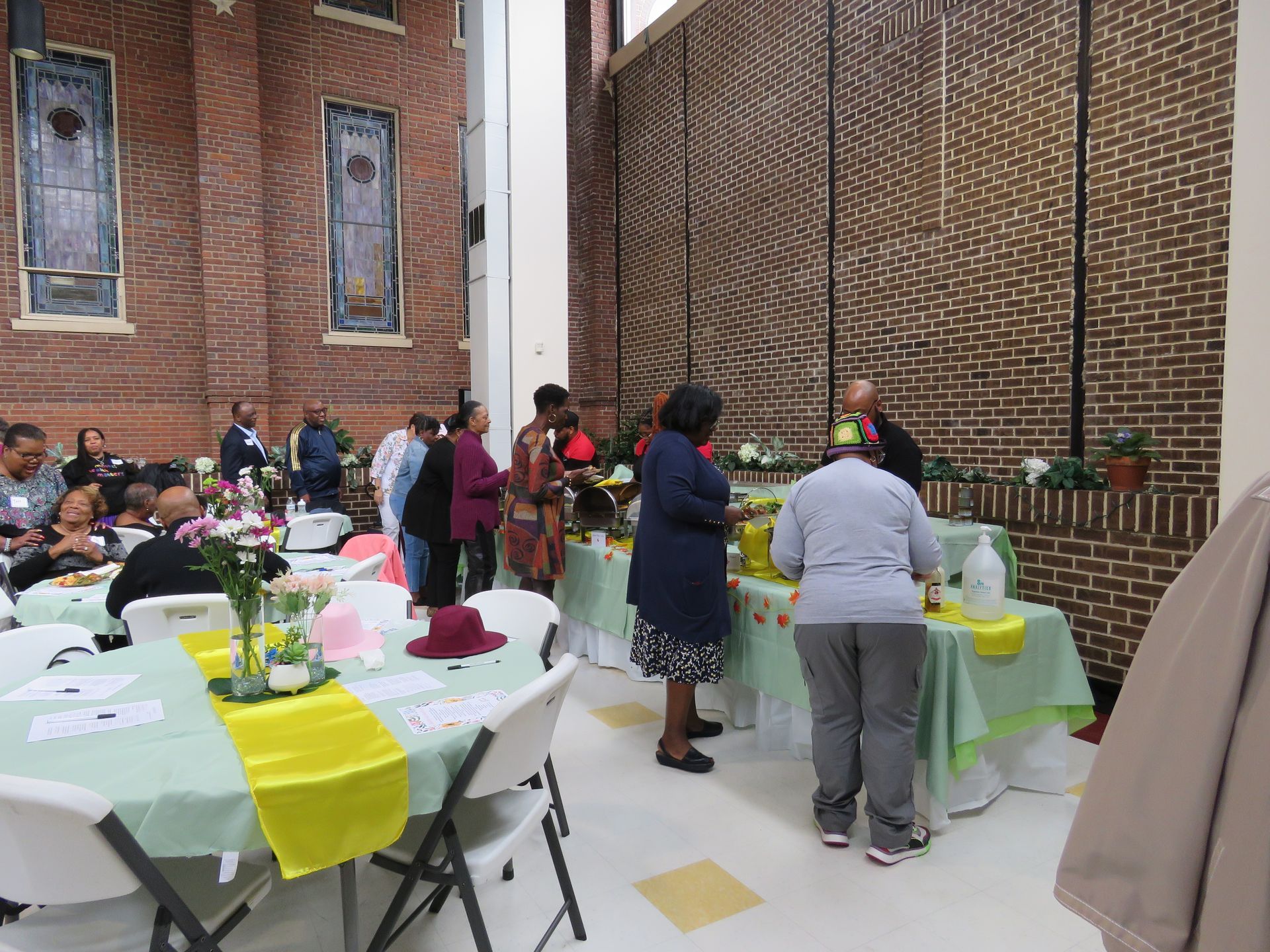 Event with tables, food, and people in a brick-walled room.  Some stand at a buffet.  Tables have decorations.