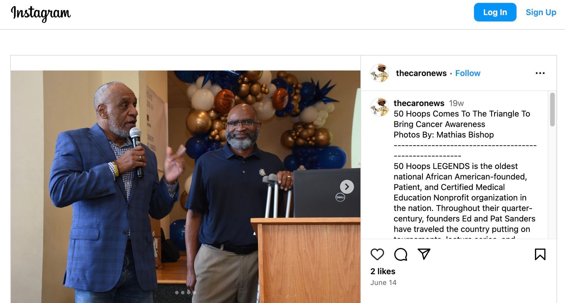 Two men speaking at a podium with a laptop, balloons in background.