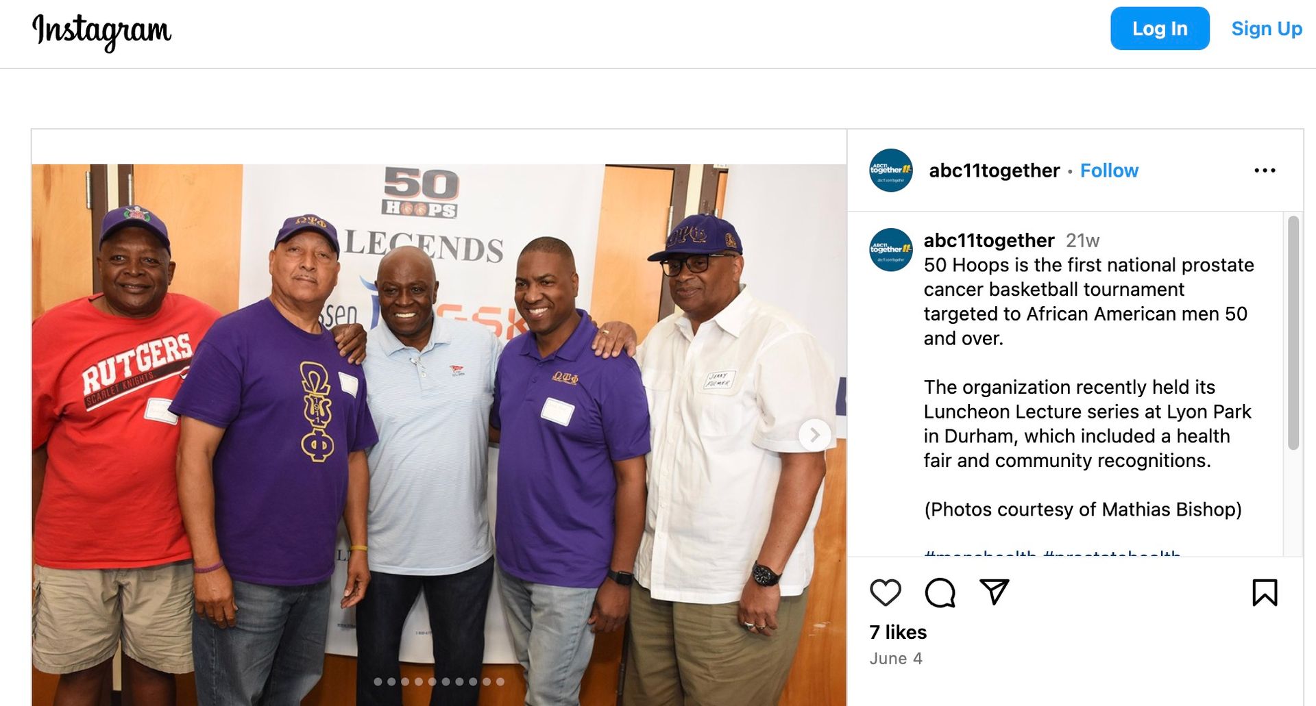 Five men pose together in front of a banner. They are smiling. One wears a red shirt, two wear purple shirts.