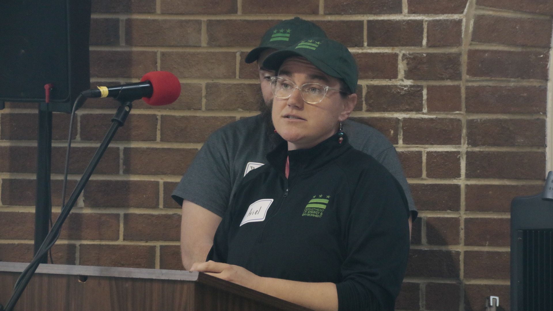 Woman speaking at podium, wearing a black shirt, glasses, and a green baseball cap. Brick wall backdrop.