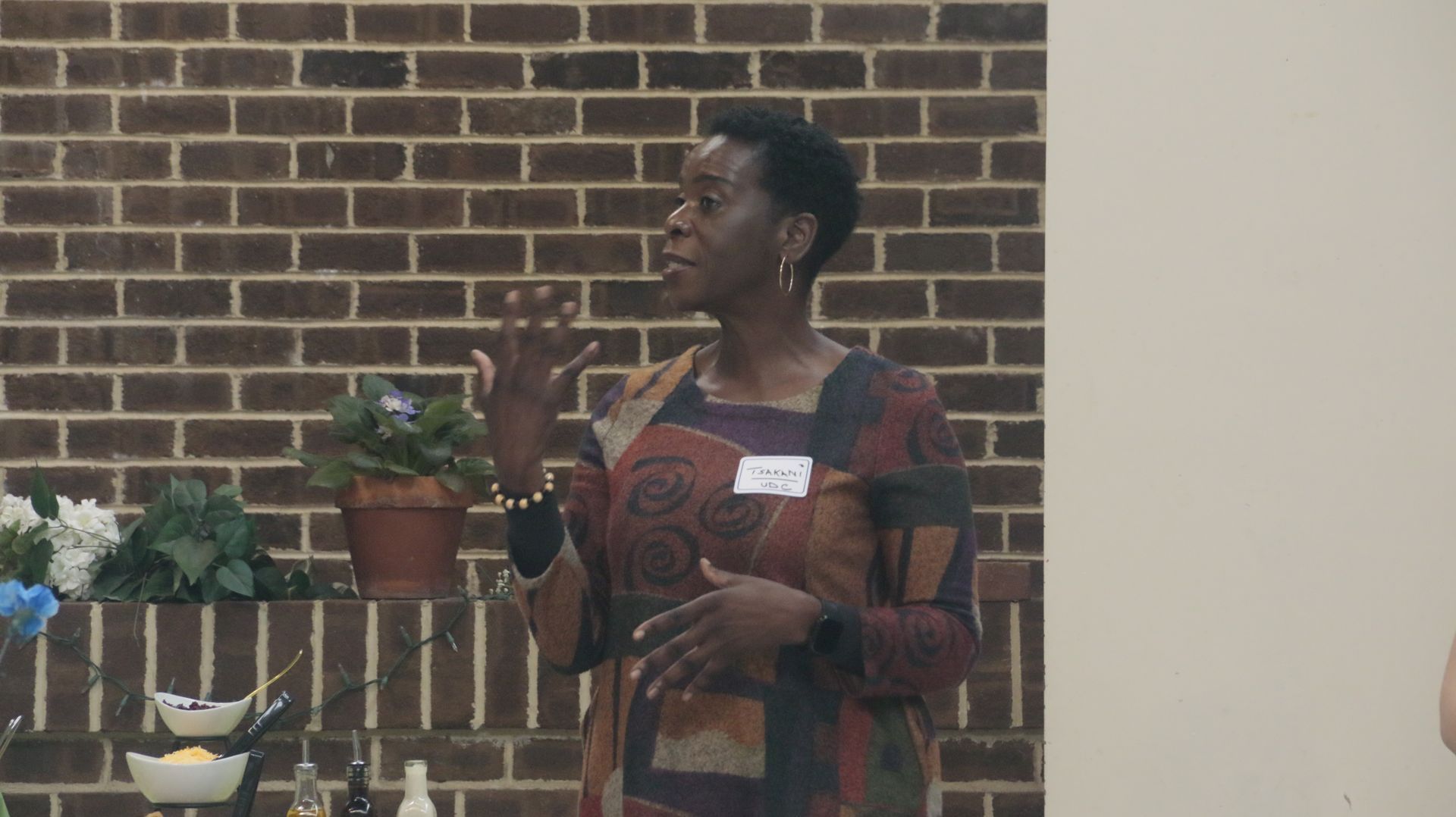 Woman speaking, gesturing with hands, in front of a brick wall and a small table.