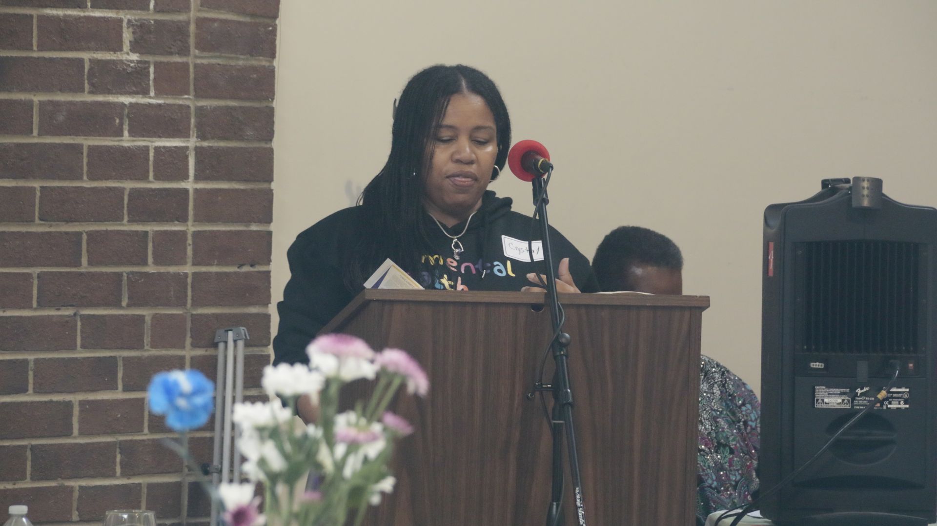 Woman speaking at a podium, flowers in the foreground, brick and beige wall backdrop.