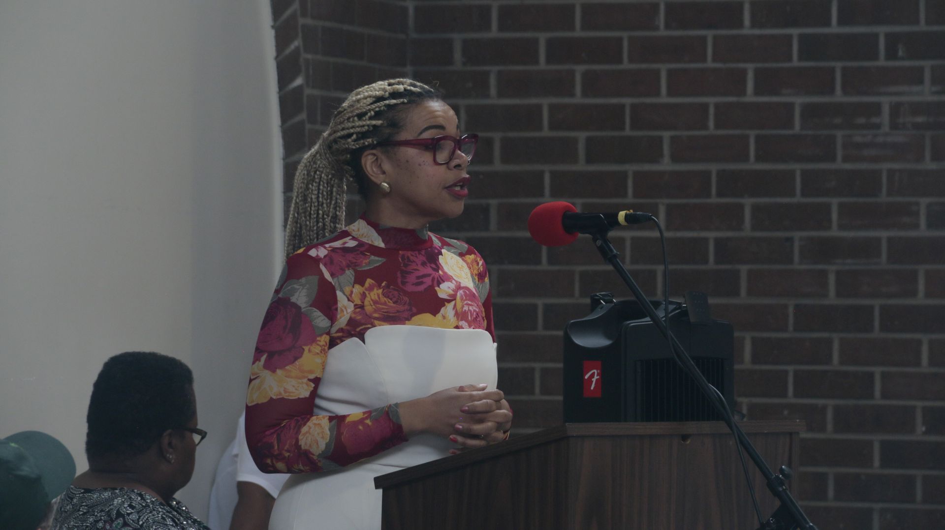 Woman in floral top speaks at a podium; brick wall in background.