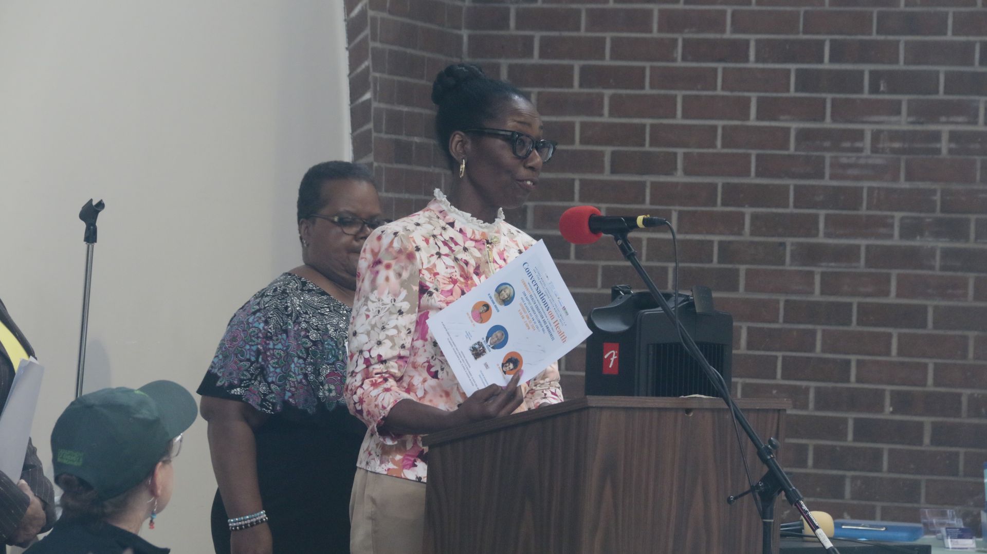 Woman speaking at a podium, holding a document, in front of a brick wall. Another woman stands behind her.