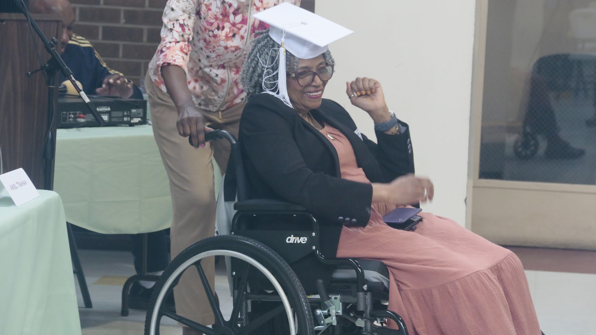 Woman in graduation cap and gown in wheelchair, smiles as pushed indoors.