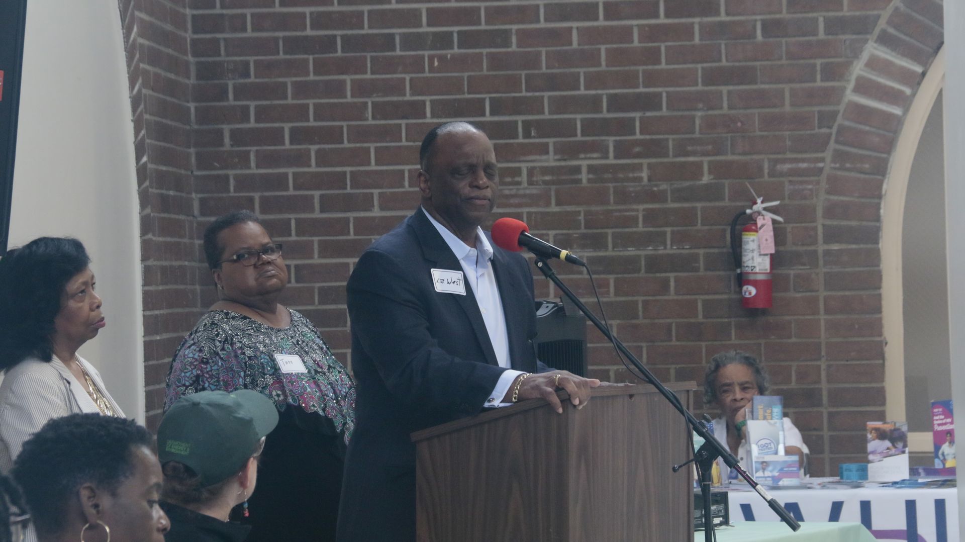 Man speaking at a podium with two women behind him and other people in the background, brick wall.