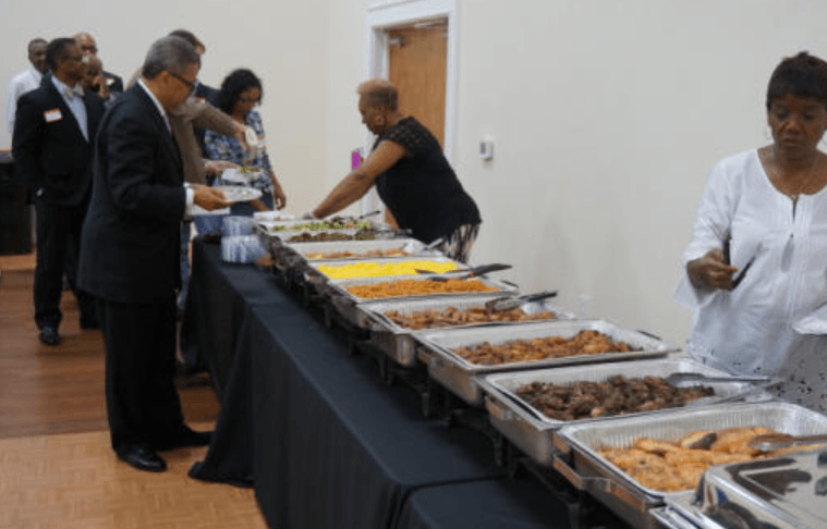People at a buffet table, selecting food from various heated serving trays in a brightly lit room.