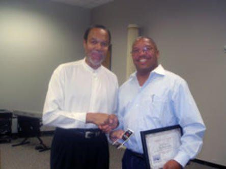 Two men shaking hands, one holding a framed document, in an office. Both men are smiling.