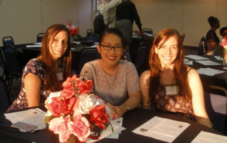 Three women at a table with flowers, smiling. Indoors, documents on table, and other people in background.
