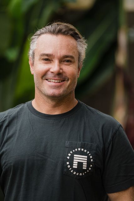 Smiling man in a dark T-shirt standing outdoors with a blurred green background