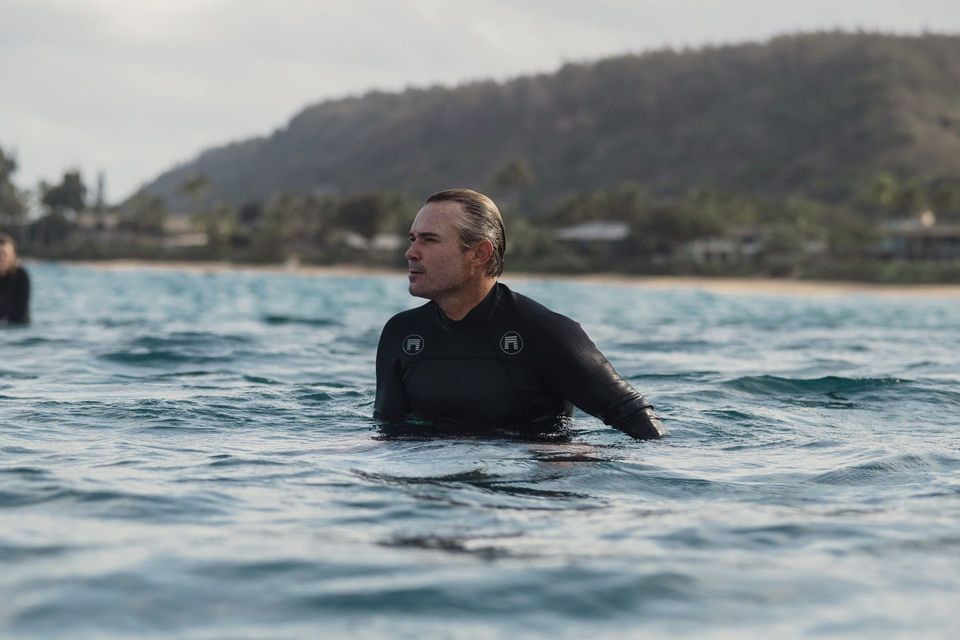 Person in a black wetsuit floating in the ocean with a shoreline and hills in the background
