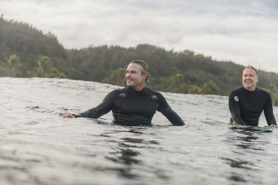Two swimmers in black wetsuits in open water, with forested hills in the background.