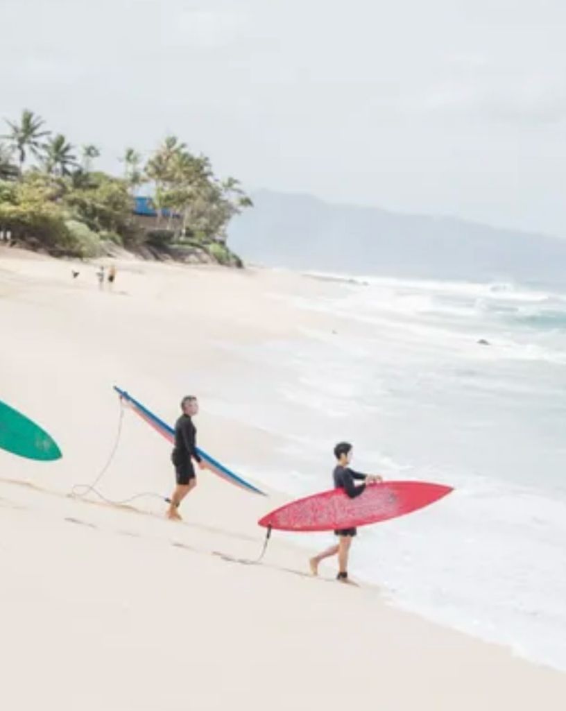 Two surfers carrying red and blue boards on a sunny beach with waves in the background