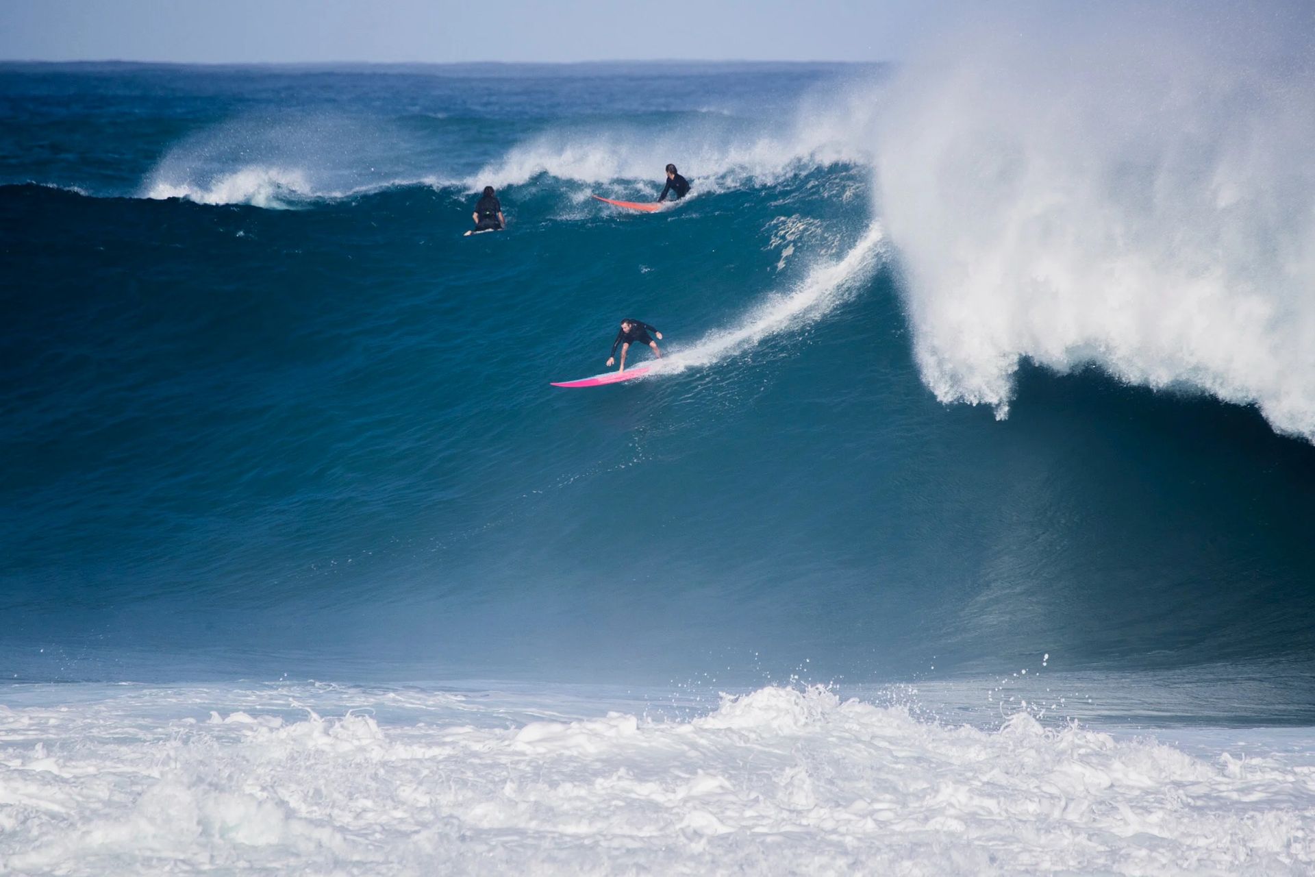 Surfer riding a pink board on a huge blue wave as others wait in the background