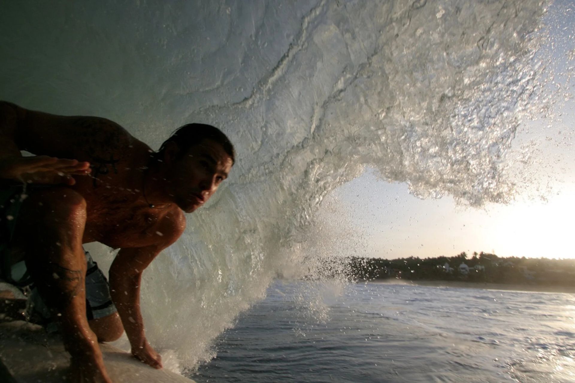 Surfer crouching inside a breaking wave at sunset, spray arcing over the water