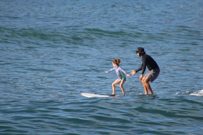 Person and child surfing on a small white board in calm blue water.