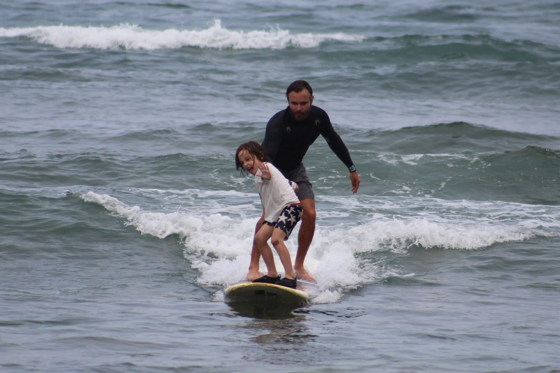 Two people surfing on a small wave in the ocean, one helping the other balance on a yellow board