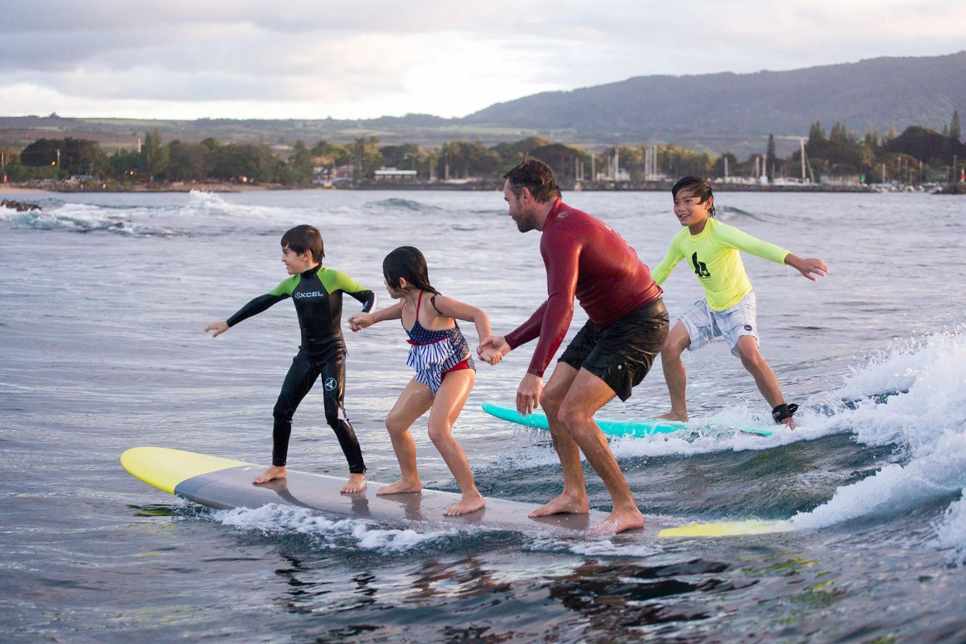 Group surfing on a small wave near shore, with two adults and two children balancing on boards
