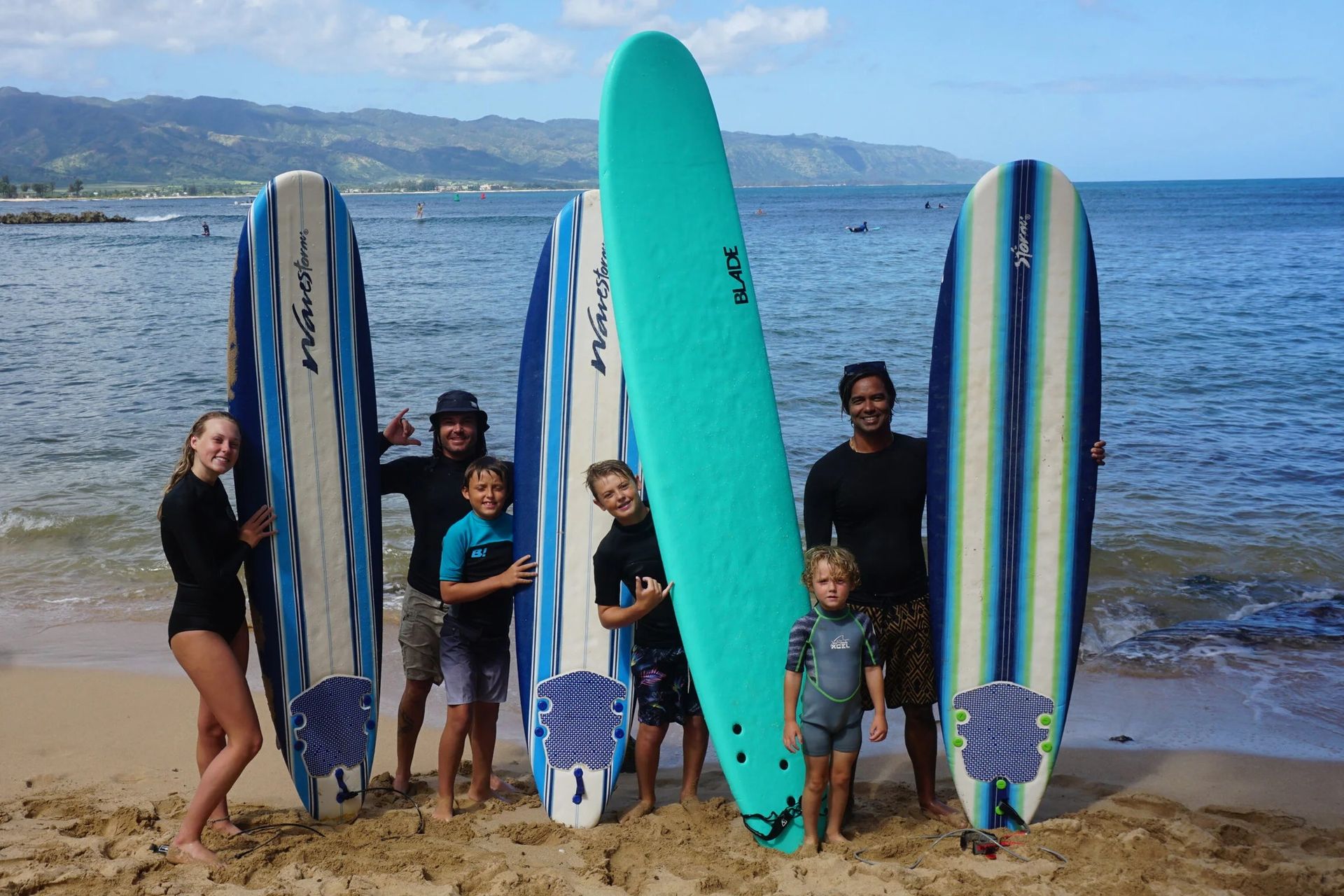 Group on a beach holding surfboards, with ocean and mountains in the background.