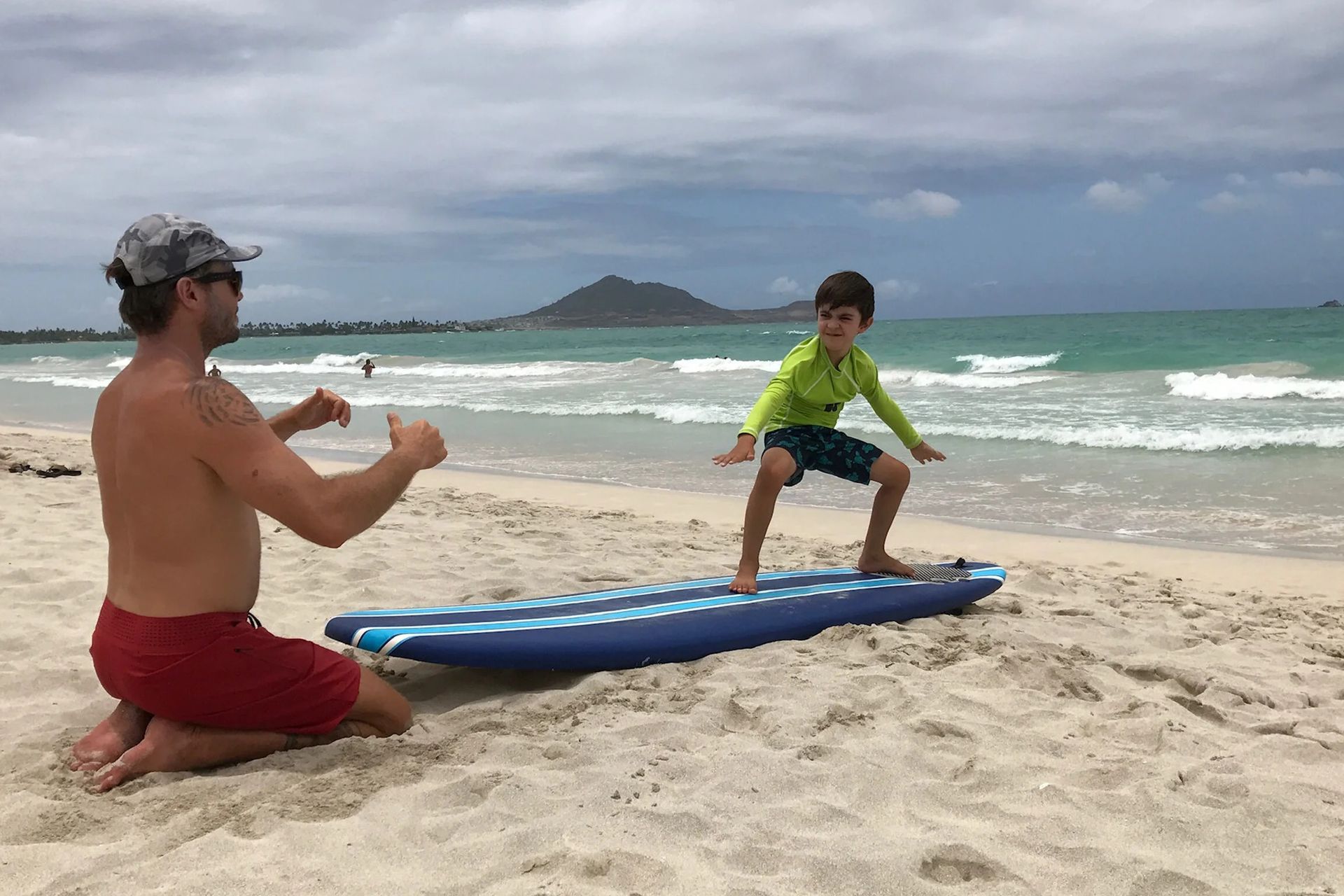 Man kneels on a sandy beach filming a child balancing on a blue surfboard by the ocean.