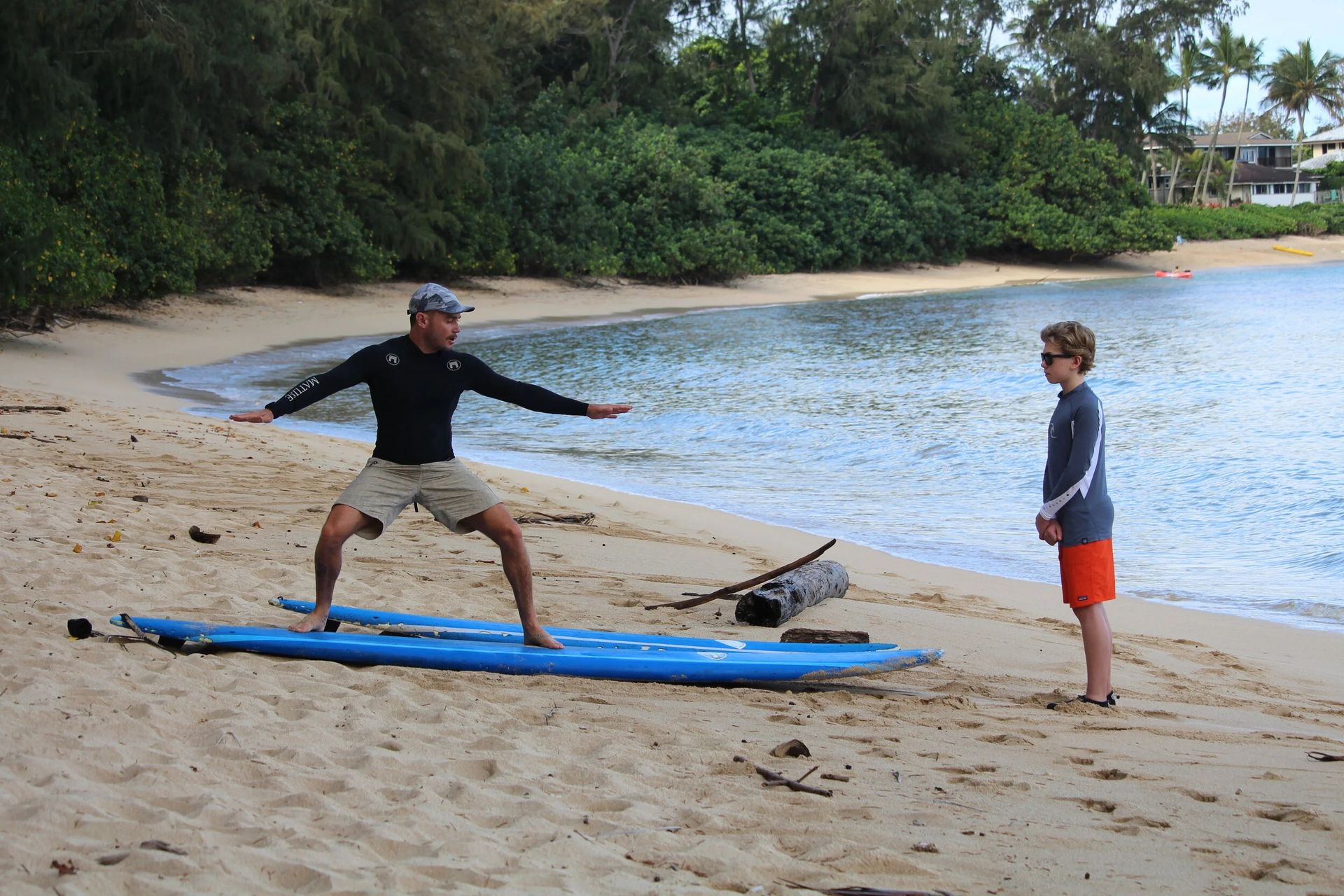 Two people on a sandy beach beside a blue kayak, one balancing on it with arms outstretched.