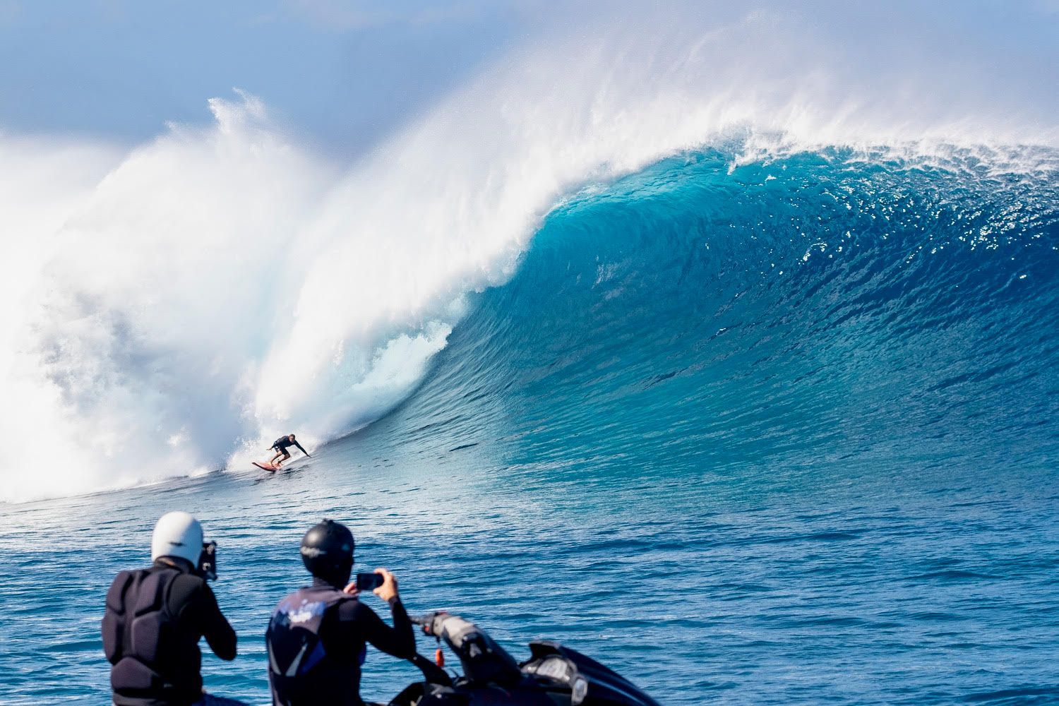 Surfer riding a large blue wave while two people watch from a boat in the foreground