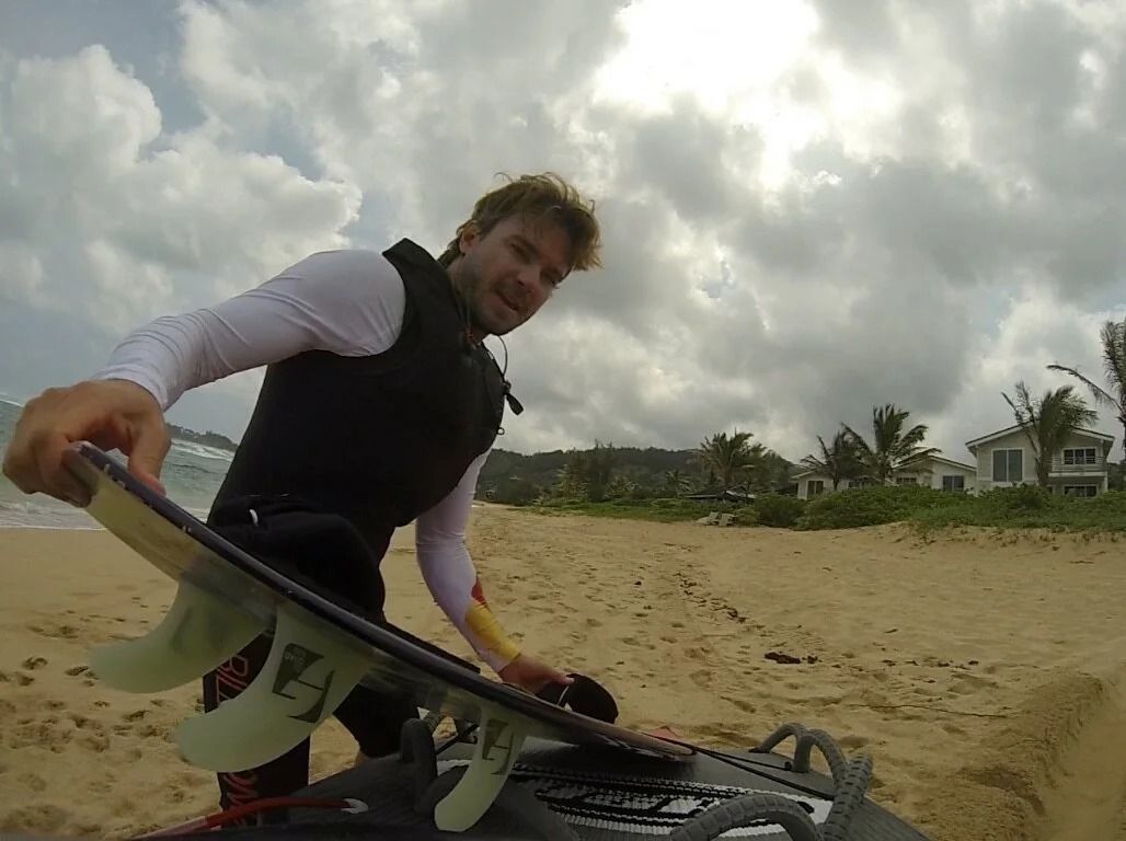 Surfer in a black vest holding a surfboard on a sandy beach under cloudy skies