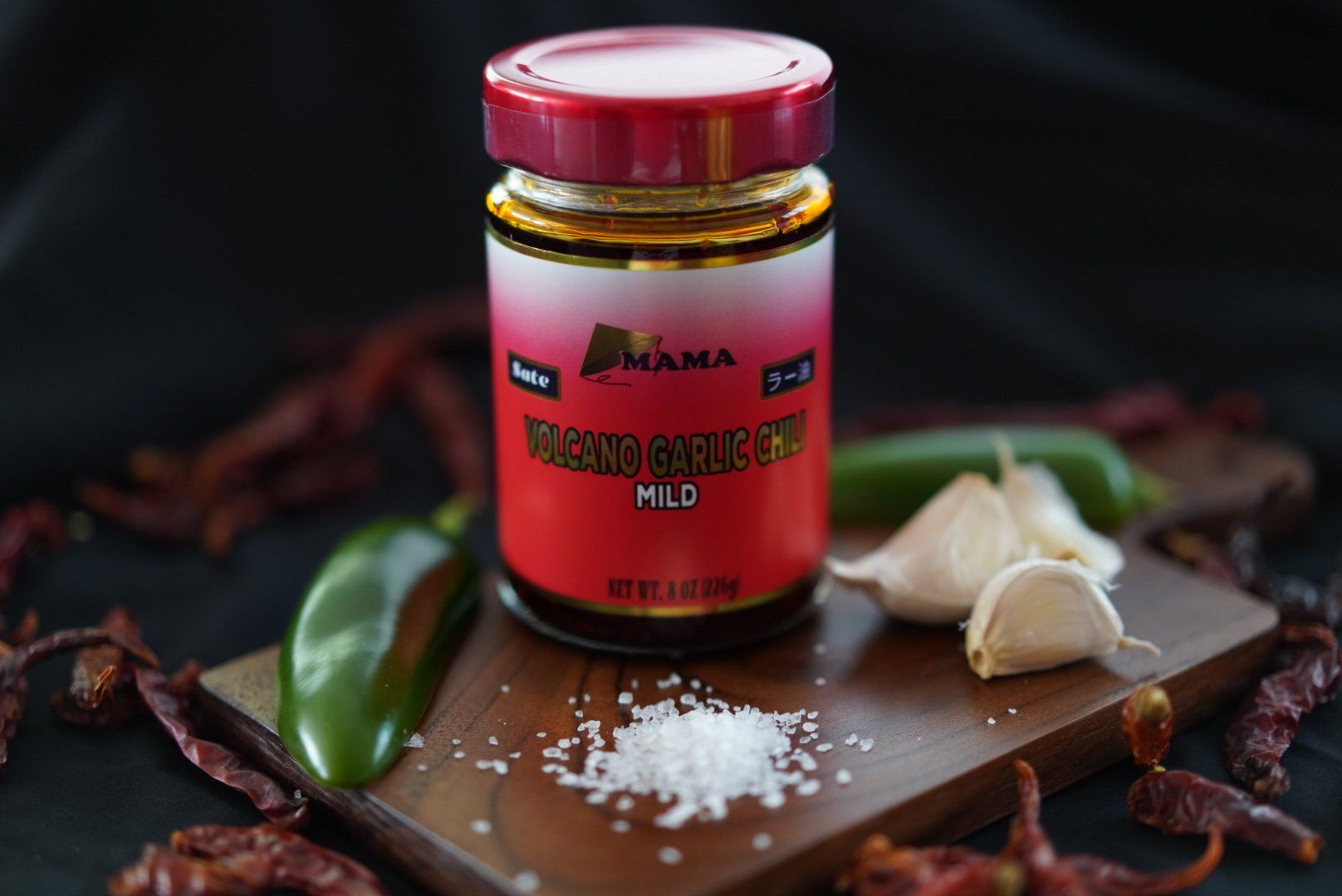 A jar of sauce is sitting on a wooden cutting board surrounded by peppers and garlic.