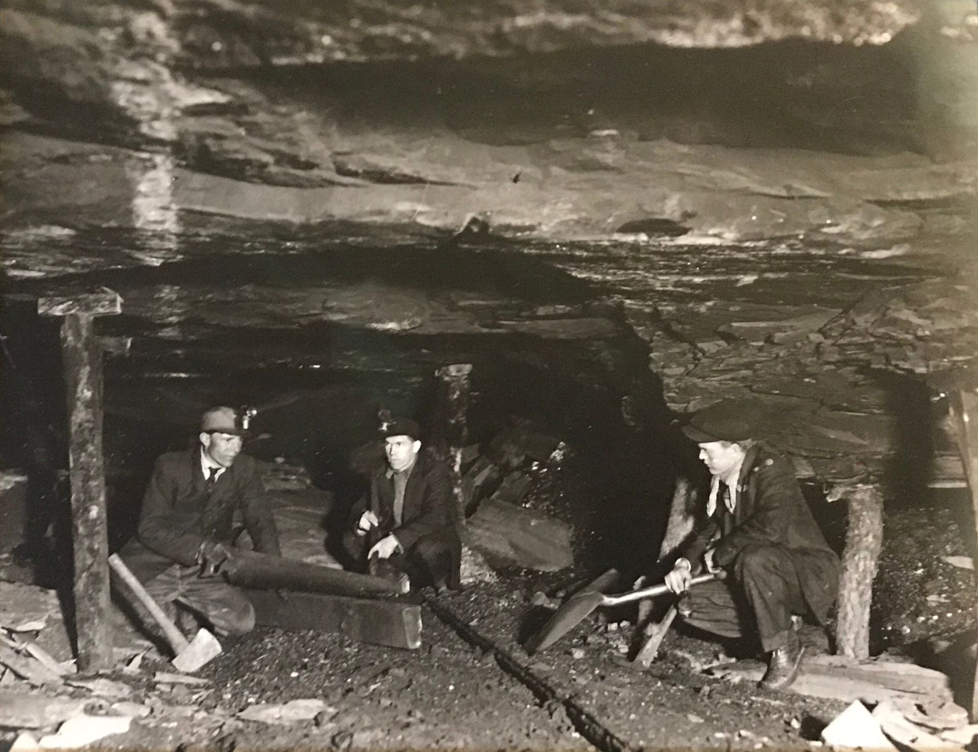 Three coal miners inside a dark mine, digging and seated with tools. Men in suits and hats, working in a cramped space.
