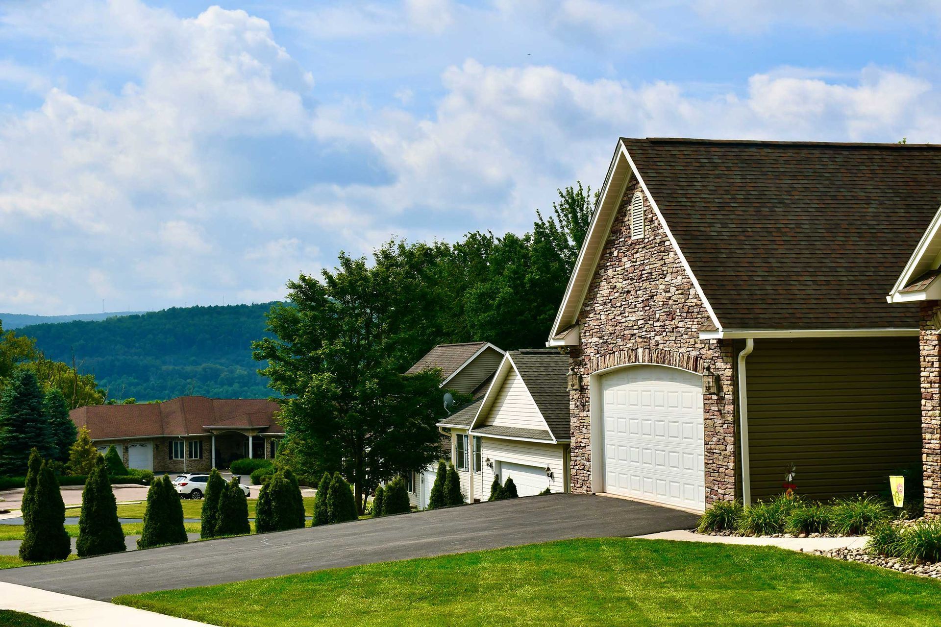 Suburban homes with a driveway on a grassy lawn, trees, and a mountain backdrop under a cloudy sky.