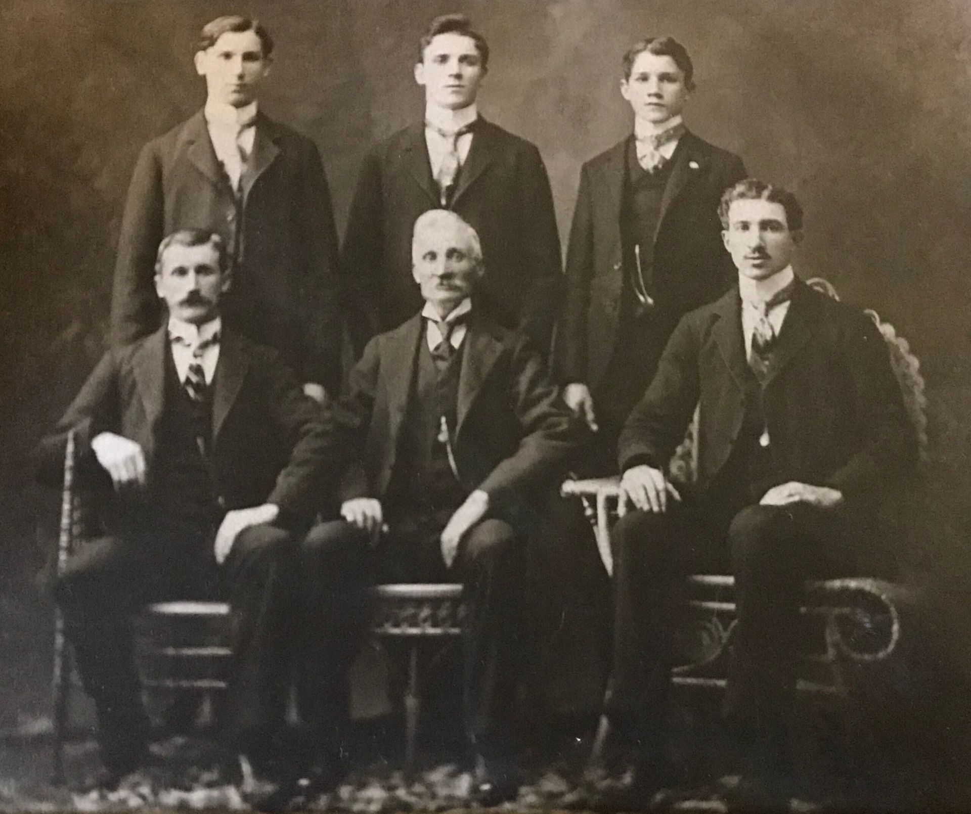 A sepia-toned portrait of six men in suits. One older man sits centered, surrounded by younger men, all formally dressed.