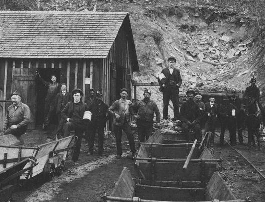 Group of coal miners standing outside a wooden structure, with mine carts visible. Some are holding containers.