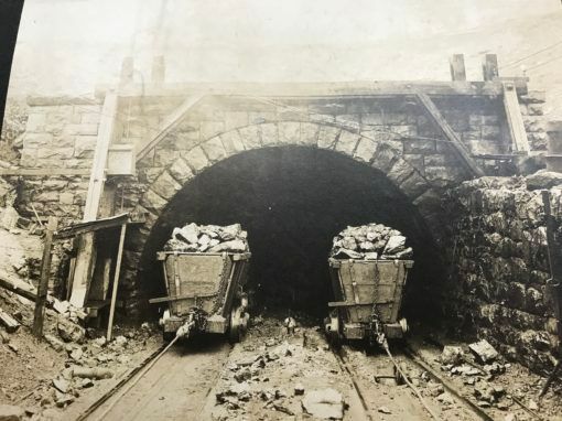 Two mine carts loaded with rocks emerge from a tunnel entrance, set in a stone arch. The setting appears old.