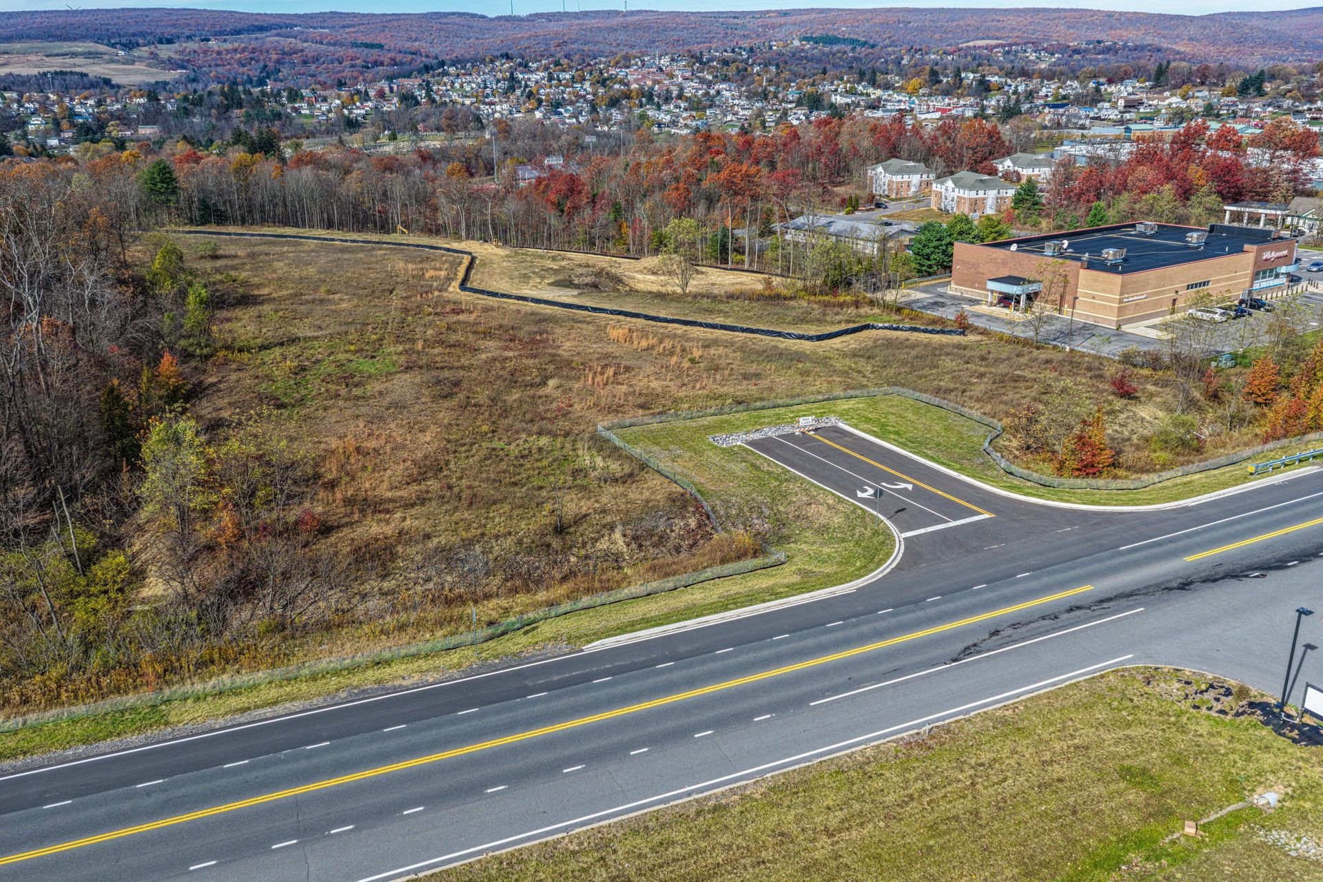 Aerial view of a vacant lot next to a road, with trees and buildings in the background.